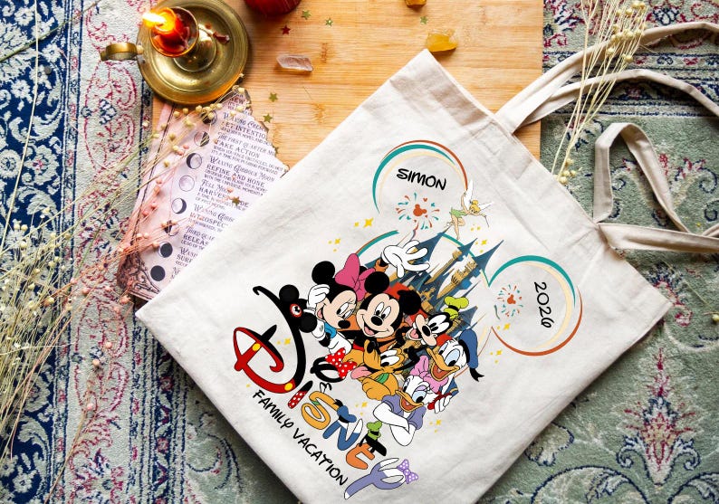 K&ouml;nnte beinhalten: Eine cremefarbene Tragetasche mit einem Disney-Familienurlaubsdesign. Das Design umfasst Micky und Minnie Maus, Donald und Daisy Duck und Goofy. Die Tasche tr&auml;gt den Text "Disney Family Vacation", den Namen "Simon" und das Jahr "2026".