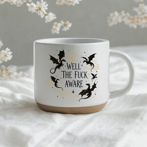 May include: White ceramic mug with a brown base. The mug features a design of black dragons, stars, and the text "Well The Fuck Aware". The mug has a curved handle.
