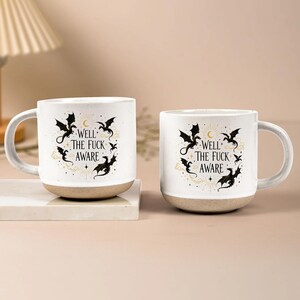 May include: Two white ceramic mugs with a beige base. Each mug features a black dragon design with the text "WELL THE FUCK AWARE" in a decorative font. The mugs have a curved handle and are set on a white surface.