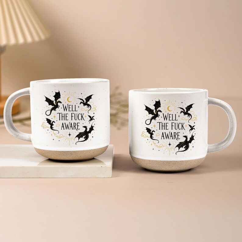 May include: Two white ceramic mugs with a beige base. Each mug features a black dragon design with the text "WELL THE FUCK AWARE" in a decorative font. The mugs have a curved handle and are set on a white surface.