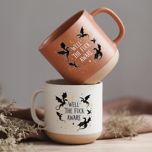 May include: Two ceramic mugs, one stacked on top of the other. The top mug is terracotta, the bottom is white. Both feature black dragon designs, a crescent moon, stars, and the text "WELL THE FUCK AWARE".