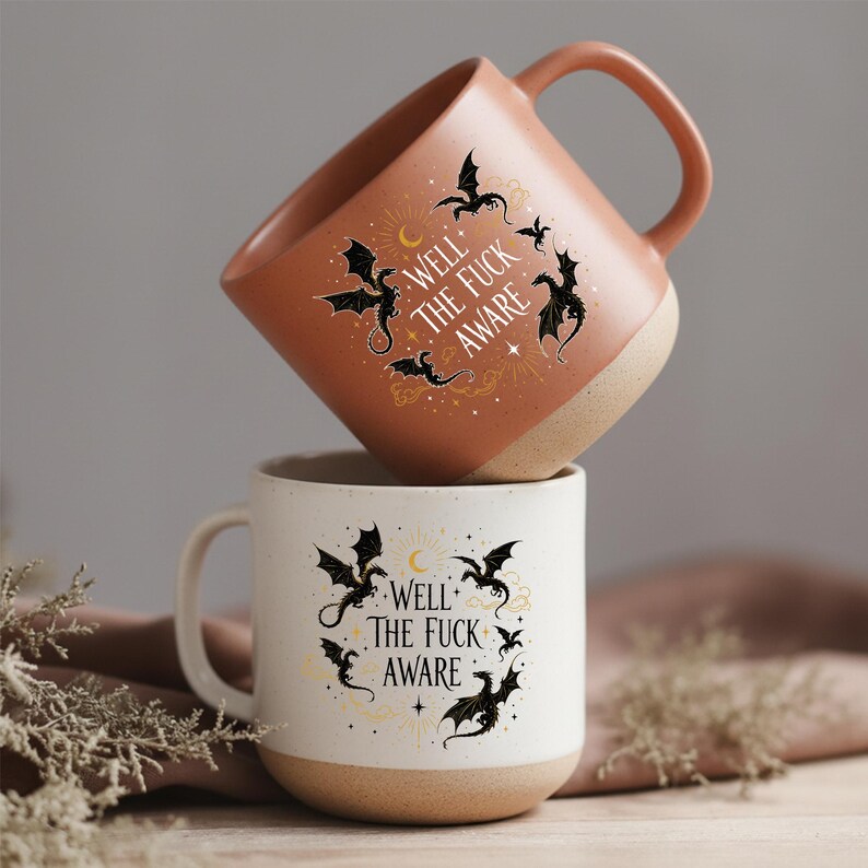 May include: Two ceramic mugs, one stacked on top of the other. The top mug is terracotta, the bottom is white. Both feature black dragon designs, a crescent moon, stars, and the text "WELL THE FUCK AWARE".
