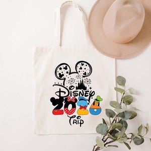 Puede incluir: Un bolso tote de color crema con un dise&ntilde;o negro y colorido de Disney 2024 Trip. El dise&ntilde;o incluye orejas de Mickey Mouse, fuegos artificiales, un castillo y los n&uacute;meros 2024 en rojo, azul y amarillo. Un sombrero beige descansa sobre la bolsa.