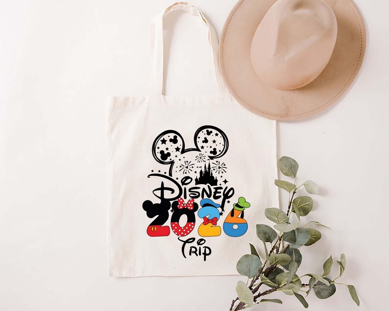 Puede incluir: Un bolso tote de color crema con un dise&ntilde;o negro y colorido de Disney 2024 Trip. El dise&ntilde;o incluye orejas de Mickey Mouse, fuegos artificiales, un castillo y los n&uacute;meros 2024 en rojo, azul y amarillo. Un sombrero beige descansa sobre la bolsa.
