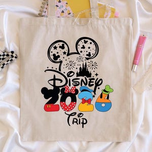 Puede incluir: Un bolso tote beige con un dise&ntilde;o de Disney 2020 Trip. El dise&ntilde;o presenta orejas de Mickey Mouse, fuegos artificiales, un castillo y los n&uacute;meros 2020 con Minnie, Donald y Goofy. Tambi&eacute;n se ven una pinza para el pelo en blanco y negro, un tubo de brillo labial rosa y un objeto de pl&aacute;stico transparente.