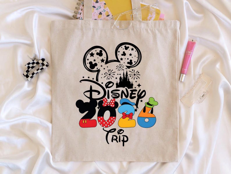 Puede incluir: Un bolso tote beige con un dise&ntilde;o de Disney 2020 Trip. El dise&ntilde;o presenta orejas de Mickey Mouse, fuegos artificiales, un castillo y los n&uacute;meros 2020 con Minnie, Donald y Goofy. Tambi&eacute;n se ven una pinza para el pelo en blanco y negro, un tubo de brillo labial rosa y un objeto de pl&aacute;stico transparente.