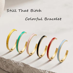 Puede incluir: Seis pulseras abiertas de metal dorado con esmalte de colores en amarillo, turquesa, rosa, negro, rojo y azul claro. El texto "Still That Bitch" y "Colorful Bracelet" está impreso en negro sobre las piezas de joyería.
