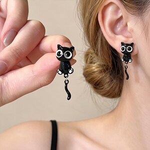 Pode incluir: Um par de brincos de gato preto com acabamento brilhante, olhos brancos grandes e detalhes de patas brancas. Cada brinco consiste numa figura de gato sentado com uma cauda curva pendente presa por um pequeno anel de metal prateado.