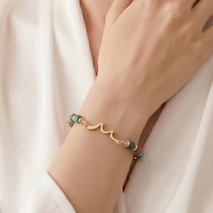 Pode incluir: Pulseira dourada com design de ondas e contas verdes. A pulseira é usada no pulso, mostrando seu design delicado e contraste de cores. O design da pulseira é simples e elegante.