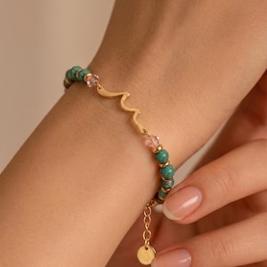 Könnte beinhalten: Goldfarbenes Armband mit türkisfarbenen Perlen, rosa Kristallen und einem wellenförmigen Golddesign. Das Armband ist mit einem kleinen goldenen Anhänger versehen. Der Schmuck wird an einem Handgelenk gezeigt.