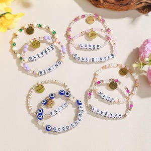 Op de afbeelding: Een verzameling kralenarmbanden in verschillende kleuren, met bedels en letterkralen. De armbanden bevatten zinnen als "Blessed", "Be Kind", "Faith" en "Good Energy". Sommige armbanden hebben goudkleurige bedels met woorden als "Lover" en "Mama".
