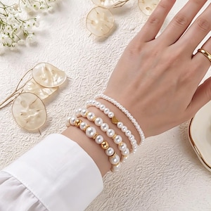 May include: A collection of pearl bracelets in various styles. Some bracelets feature gold accents and a small gold heart charm. The pearls are white and the bracelets are worn on a wrist with a gold ring.
