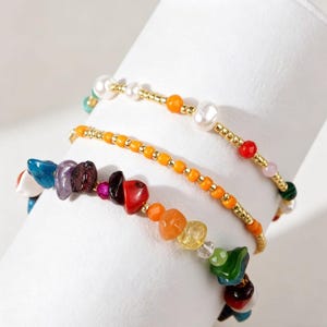 May include: Three colorful beaded bracelets. One bracelet features a mix of colorful gemstones. Another has orange beads with gold accents. The third bracelet has white pearls and gold accents. The bracelets are displayed on a white surface.