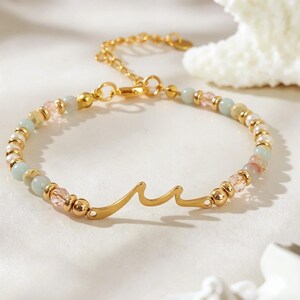 May include: A gold-toned bracelet featuring a central wave-shaped metal charm, accented with alternating light blue, pale pink, and metallic gold beads. The piece includes a delicate gold-toned chain with a lobster clasp closure for adjustment.