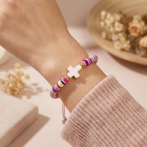 Könnte beinhalten: Ein farbenfrohes Perlenarmband mit einem weißen Kreuzanhänger. Das Armband hat eine Mischung aus regenbogenfarbenen Perlen und goldenen Akzenten. Das Armband befindet sich am Handgelenk einer Person.