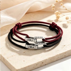 Puede incluir: Dos pulseras de cordón ajustable, una burdeos y otra negra, cada una con un eslabón de metal plateado grabado con la fecha 05.20.2025 e iniciales separadas por un corazón, A corazón J en una y J corazón A en la otra.