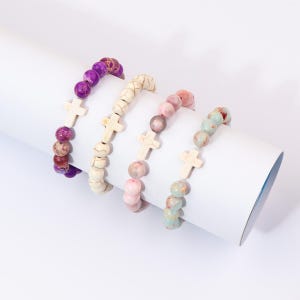 Puede incluir: Cuatro pulseras de cuentas con dijes de cruz. Las pulseras presentan una variedad de colores, incluyendo morado, crema, rosa y azul-verdoso. Cada pulsera tiene un dije de cruz. Las cuentas son de piedra.