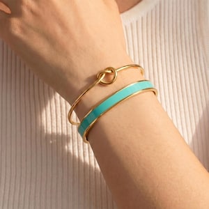 May include: A set of two gold-toned bracelets. One is a thin, flexible wire bangle featuring a central knot detail. The second is a wider, rigid cuff bracelet with a smooth, vibrant turquoise enamel inlay that spans the entire outer surface.