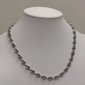 May include: A silver-toned chain necklace featuring a series of uniform, oval-shaped links with a central bar detail in each link. The necklace is displayed on a white neck form, showcasing the polished metallic finish and the consistent design.