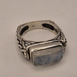 May include: A silver ring featuring a rectangular, translucent moonstone cabochon set in a band with an intricate, woven-style metal texture. The metal has darkened, recessed areas that highlight the raised, braided pattern along the band.