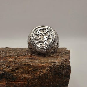 May include: A silver-toned ring featuring a circular face with an intricate, raised cutout pattern of swirling, organic shapes. The band of the ring displays a detailed, openwork design that extends around the sides, showcasing a metallic finish.