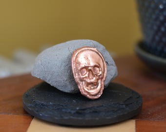 Puede incluir: Un pequeño emblema de calavera de color cobre metálico con un relieve detallado de un cráneo humano. La calavera presenta cuencas oculares huecas, una cavidad nasal y una mandíbula abierta, apoyada contra una piedra gris redondeada.