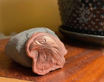 Puede incluir: Un emblema de metal color cobre con una escultura en relieve de una cabeza de águila de perfil, apoyada contra una piedra gris, texturizada e irregular. El diseño del águila muestra plumas detalladas y un pico afilado con acabado metálico.