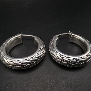 May include: A pair of silver hoop earrings featuring a thick, rounded profile with a textured, braided pattern across the surface. The earrings have a polished metallic finish and a hinged post closure mechanism visible on the inner side.