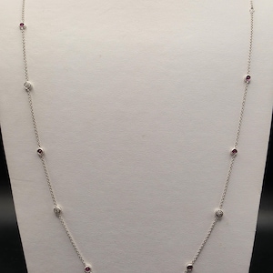 May include: A long silver chain necklace featuring alternating bezel-set round red gemstones and small round white diamonds. The delicate metal chain links connect each stone station, creating a spaced pattern along the entire length of the jewelry piece.