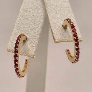 May include: A pair of gold hoop earrings featuring a row of small, round, deep red rubies set along the front-facing curve of each hoop. The earrings have a polished gold finish and a classic open-circle design, showcasing a vibrant contrast between the metallic gold and the dark red gemstones.