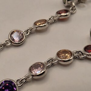 May include: A pair of silver-toned drop earrings featuring a vertical chain of bezel-set, round-cut multicolored gemstones. The stones include shades of purple, light pink, champagne, and deep red, with a green stone at the top post.