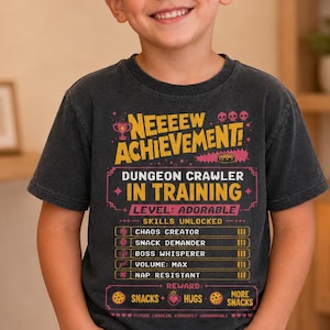 May include: A charcoal gray short-sleeve t-shirt featuring a graphic print with the text NEEEEW ACHIEVEMENT! DUNGEON CRAWLER IN TRAINING. LEVEL: ADORABLE. SKILLS UNLOCKED: CHAOS CREATOR, SNACK DEMANDER, BOSS WHISPERER, VOLUME: MAX, NAP RESISTANT.