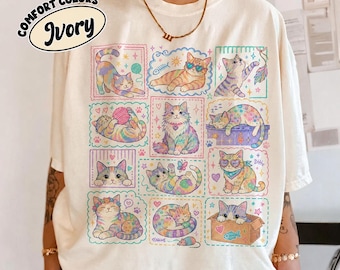 Puede incluir: Una camiseta de color marfil con una cuadrícula de doce ilustraciones coloridas de gatos en varias posturas, incluyendo gatos jugando con lana, usando gafas de sol y sentados en una caja. El gráfico incluye pequeñas estrellas, corazones y espinas de pescado.