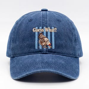 May include: A denim blue baseball cap with the phrase "Guess What?" above a graphic of a chicken. The chicken is brown, white, and red, set against blue vertical stripes. The cap has a curved brim and a button on top.