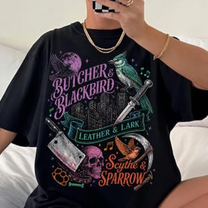 May include: Black t-shirt with a graphic design featuring a bird, a knife, a skull, and the words "Butcher & Blackbird", "Leather & Lark", and "Scythe & Sparrow". The design includes a cityscape, musical notes, and a crescent moon.