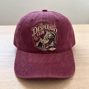 May include: A maroon baseball cap with a curved brim. The cap features the text "The Desperado Club" above an illustration of a cat wearing glasses and holding a cocktail. The phrase "So Fun It Hurts" is also included.