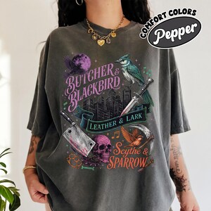 May include: Dark gray t-shirt with a graphic design featuring a purple moon, a blue bird, a knife, a cleaver, a skull, and the text "BUTCHER & BLACKBIRD LEATHER & LARK SCYTHE & SPARROW". The shirt has the text "COMFORT COLORS Pepper".