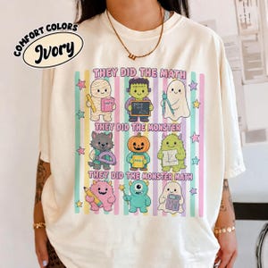 May include: An ivory t-shirt featuring a graphic print of nine cartoon monsters engaged in math-related activities. The text reads "THEY DID THE MATH," "THEY DID THE MONSTER," and "THEY DID THE MONSTER MATH" over vertical pastel stripes and stars.