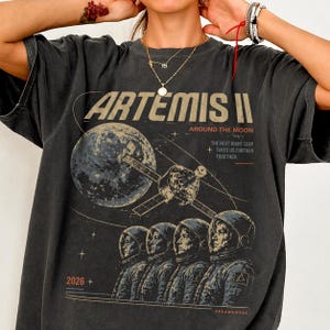May include: Dark gray t-shirt featuring a graphic design for the Artemis II mission. The design includes the text "ARTEMIS II", a depiction of the moon, a spacecraft, and astronauts. The shirt also has the text "AROUND THE MOON", "THE NEXT GIANT LEAP + TAKES US FURTHER TOGETHER", and "2026 +".