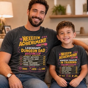 May include: Two charcoal gray t-shirts featuring colorful graphic prints. The adult shirt reads "NEEEEEW ACHIEVEMENT! LEGENDARY DUNGEON DAD" with skill lists. The child shirt reads "NEEEEEW ACHIEVEMENT! DUNGEON CRAWLER IN TRAINING" with skill lists.