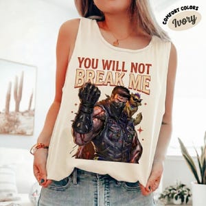 May include: Ivory-colored tank top with the text "YOU WILL NOT BREAK ME" and a graphic of a muscular figure with a black mask and spiked gauntlet. The graphic also includes a small animal with sunglasses. The words "Comfort Colors" and "Ivory" are also visible.