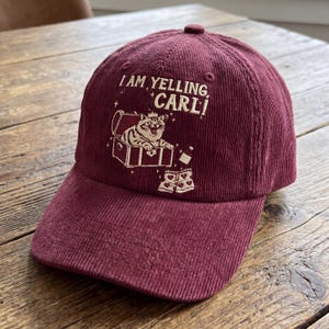 May include: A burgundy corduroy baseball cap with the embroidered phrase "I AM YELLING, CARL!" above a cartoon cat in a treasure chest. The cap also features a small bottle and a pair of underwear. The hat has a curved brim and a button on top.