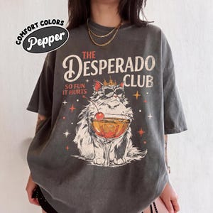 May include: Dark gray t-shirt with a graphic of a cat wearing sunglasses and a crown, holding a cocktail. The text reads "THE DESPERADO CLUB" and "SO FUN IT HURTS". The shirt has a vintage, distressed look.
