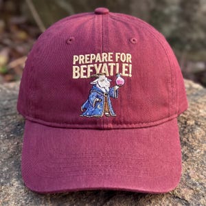 May include: A maroon baseball cap with the embroidered phrase "PREPARE FOR BEFVAT!E!". The cap features an embroidered illustration of a goat in a wizard's robe holding a potion. The cap is made of a soft, durable material.