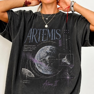 May include: Dark gray t-shirt featuring the word "ARTEMIS" in a stylized font, with a graphic of a planet and spacecraft. The shirt also includes text about the Orion spacecraft and SLS launch system. The coordinates 28.5383° N 80.6042° W are printed on the shirt.