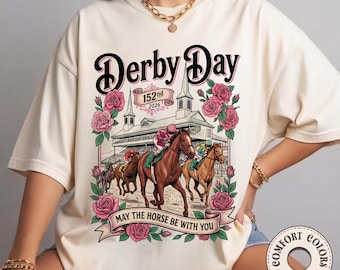 May include: Ivory-colored t-shirt with a graphic featuring a horse race scene. The design includes the text "Derby Day" in a black script, the number "152nd 2026", and the phrase "May the horse be with you". Pink roses frame the scene.