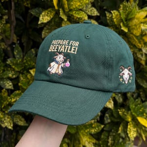 May include: A dark green baseball cap with the embroidered text "PREPARE FOR BEEYATLE!". The cap features an embroidered cartoon goat holding a potion and a goat head on the side. The cap is made of a durable fabric.