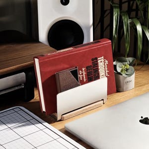 May include: A white and wood-toned desk organizer holds a red book with white text and a small, metallic card holder. A small potted plant with the word "cute" on the pot sits nearby. A laptop and speaker are also visible.