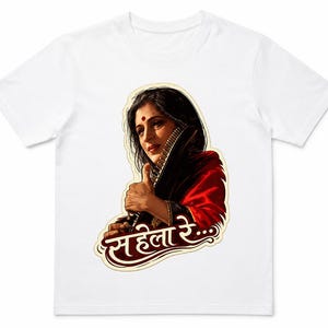 May include: A white short-sleeved t-shirt featuring a graphic print of a person with dark hair, a red bindi, and a red and black garment. The person is holding a musical instrument. Below the figure is the text Sahela Re in Devanagari script.