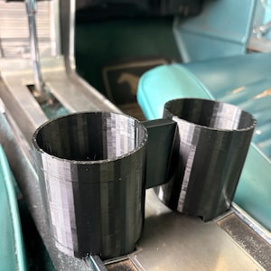 May include: Black 3D-printed car cup holder with two circular openings. The cup holder is designed to fit in a car's center console. The interior of the car has teal seats and a silver console.
